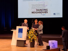 Drei Personen stehen auf einer Bühne neben einem Podium, lächeln und unterhalten sich. Auf einem Bildschirm hinter ihnen ist „Tanzende Nachbarn Dancing Neighbours“ zu sehen. Daneben steht ein Blumenarrangement und davor steht auf einem kleinen Schild „C U THE UNSEEN“.