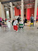 A group of elderly people sit and stand around colourful chairs in a large, bright room with high windows and red curtains. Some people are chatting while others are observing, creating a lively and sociable atmosphere.
