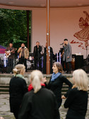 A band plays on an outdoor stage with microphones, guitar and saxophone. Several people dressed in black stand in the foreground and watch. Trees and a large mural can be seen in the background.