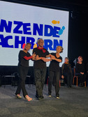 Three older adults dressed in black hold hands and dance on stage with smiles on their faces. Behind them, a large screen displays bold blue and yellow text. Other people sit in the background and watch the performance.