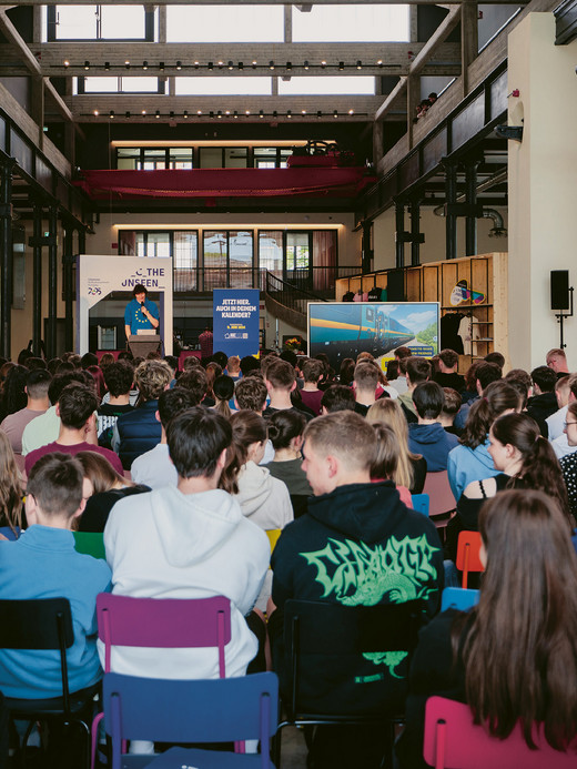 Ein großes Publikum sitzt auf bunten Stühlen in einem geräumigen, modernen Raum mit hohen Decken und freiliegenden Balken. Eine Person steht auf einer Bühne mit einer Leinwand und einem Podium vor großen Fenstern und spricht zur versammelten Menge.