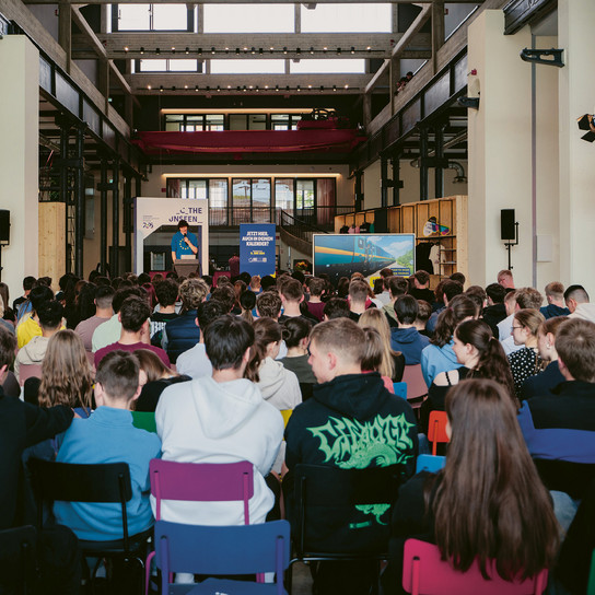 A large group of people seated indoors at an event. The room has high ceilings and a stage at the front with a speaker addressing the audience. Various banners and lights decorate the space. Attendees are facing forward, listening attentively.