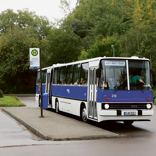 A blue and white bus is parked at a bus stop on a wet road. The route display reads "Karl-Marx-Stadt." A bus stop sign is visible, and trees are in the background. It's a cloudy day.