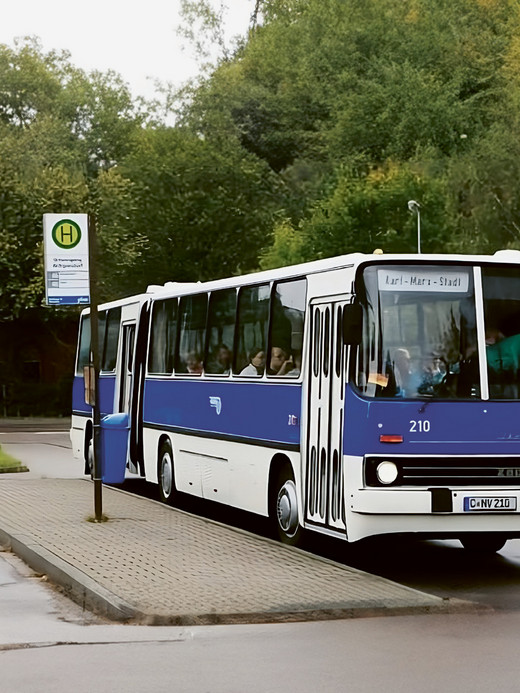 An einem regnerischen Tag steht ein blau-weißer Bus an einer Bushaltestelle. Das angezeigte Ziel lautet „Karl-Marx-Stadt“. Der Bus steht auf einem gepflasterten Platz neben einer nassen Straße, umgeben von üppigen grünen Bäumen.