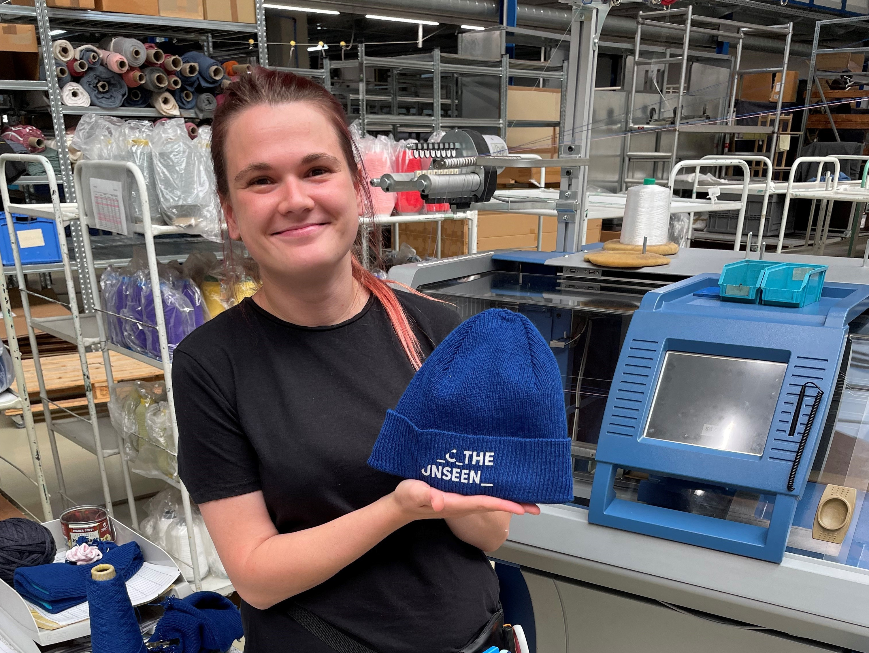 A person with a ponytail and a black shirt is holding a blue beanie in a textile factory. On the beanie is a white text with the inscription "FROM THE UNSEEN". Various machines, threads and equipment can be seen in the background.