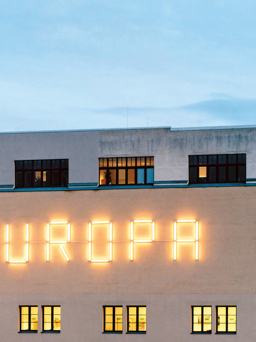 Ein Gebäude mit einer großen Neonreklame, auf der das Wort „EUROPA“ steht, vor einer hellen Fassade. Die oberen Fenster sind dunkel, während die unteren Fenster ein warmes Licht ausstrahlen. Der Himmel ist von einem sanften Blau, das den Abend andeutet.