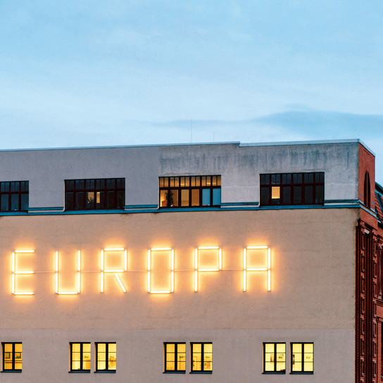 Ein Gebäude mit einer großen Neonreklame, auf der das Wort „EUROPA“ steht, vor einer hellen Fassade. Die oberen Fenster sind dunkel, während die unteren Fenster ein warmes Licht ausstrahlen. Der Himmel ist von einem sanften Blau, das den Abend andeutet.