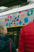 Ein bunter Zug mit geometrischen Formen an der Seite hält an einem Bahnhof. Menschen in Winterkleidung stehen daneben und wirken etwas unscharf. Die Zugtür zeigt die Nummer eins, was darauf hindeutet, dass es sich möglicherweise um den ersten Wagen handelt.