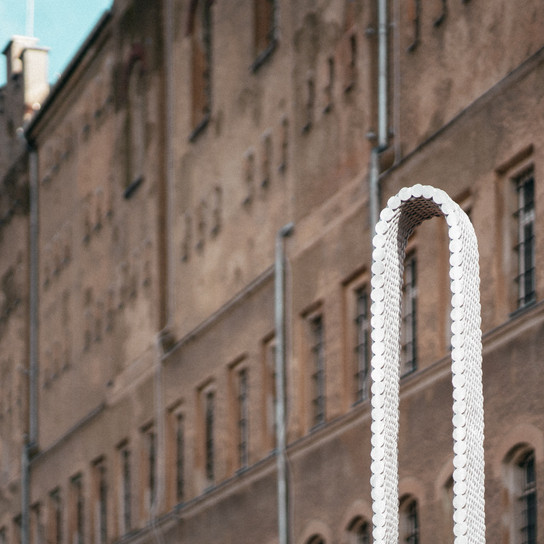 Das Bild zeigt eine Nahaufnahme eines Backsteingebäudes mit mehreren Fenstern, die teilweise von einer freistehenden modernen Metallskulptur im Vordergrund verdeckt werden. Der Himmel ist klar und blau.