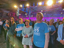 A group of people, many wearing blue "Chemnitz 2025 Volunteer" shirts, are standing in an interior room at an event. They are smiling and watching something from offstage, with colourful lighting in the background.