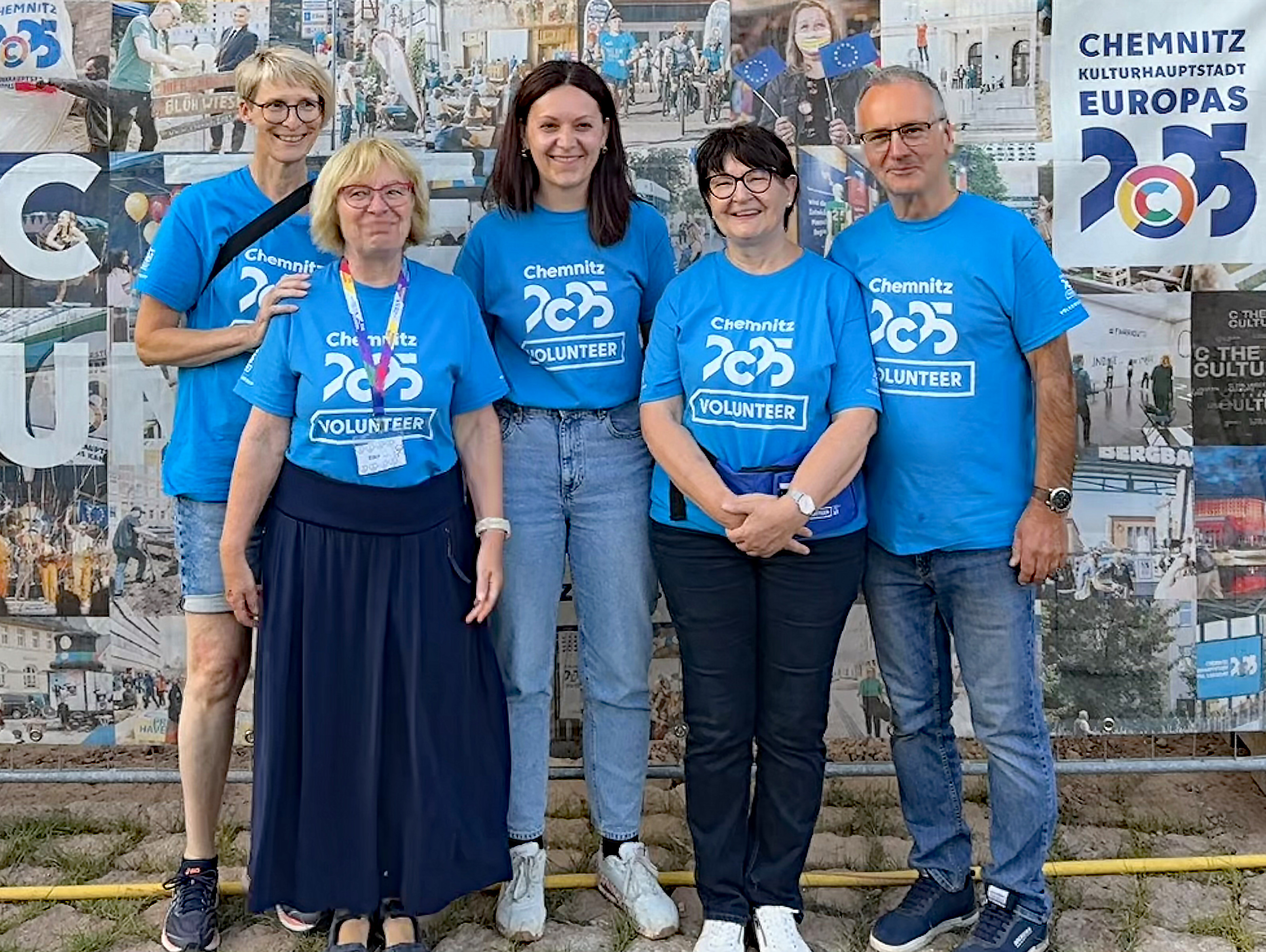 Fünf Personen stehen lächelnd vor einem Wandgemälde, alle tragen blaue "Chemnitz 2025 VOLUNTEER"-Shirts, die für Chemnitz als Kulturhauptstadt Europas 2025 werben.