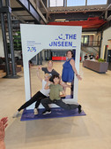 Four people pose playfully in a large photo frame labelled "THE UNSEEN" in an event hall. Two people are standing, two are crouching in front of it and smiling. The display advertises Chemnitz as European Capital of Culture 2025.