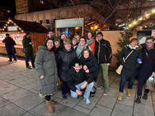 Eine Gruppe von Menschen lächelt und posiert gemeinsam auf einem Wintermarkt im Freien. Sie stehen vor einem Holzstand mit festlichen Lichtern und einem Schild mit der Aufschrift „Winterlounge“. Die Atmosphäre ist lebhaft und fröhlich.