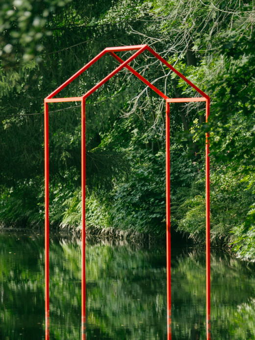 Eine rote, minimalistische Skulptur in Hausform steht inmitten eines üppigen grünen Waldes und spiegelt sich wunderschön im ruhigen Wasser darunter. Das offene Gerüst bildet einen Kontrast zum dichten umgebenden Laub.