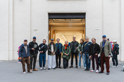 A group of eleven adults stand side by side in front of a large, open industrial gate and pose for a photo. Most of them are wearing jackets and casual clothes. The background is a plain, light-coloured wall.