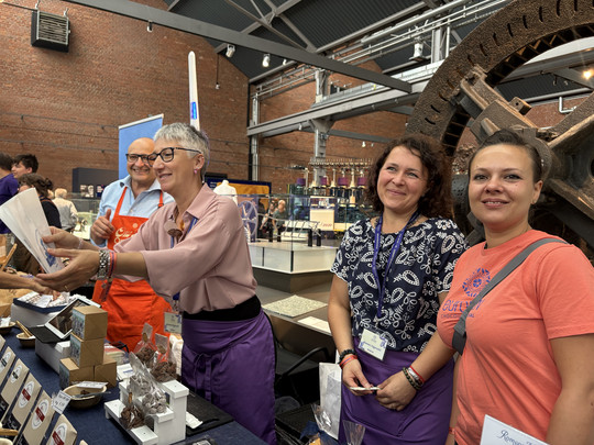 Four smiling people stand behind a table with various chocolates and treats at a market or event in a hall, with industrial machinery and brick walls in the background. A woman hands an item to a customer.