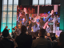 Ein Redner steht an einem Podium und spricht zu einem Publikum, während hinter ihm ein großer Bildschirm eine jubelnde Menge zeigt. Mehrere Personen im Publikum machen Fotos, und die Veranstaltung scheint in einem Innenraum stattzufinden.