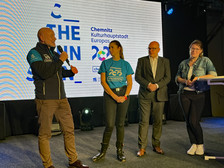 Four people stand on a stage and speak to each other. One man is holding a microphone, another man in a suit is standing next to him, a woman in a blue volunteer shirt and another woman with glasses are listening. Behind them is a banner reading "Chemnitz European Capital of Culture 2025".