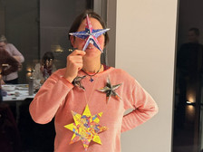 Eine Person, die einen rosafarbenen Pullover trägt, hält einen blauen sternförmigen Schmuck vor ihr Gesicht, wobei zwei weitere bunte Sternornamente an ihrem Hals hängen. Im Hintergrund sind weitere Personen zu sehen.