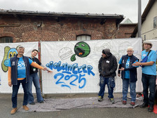 Five people stand in front of a graffiti mural with the words "Volunteer 2025" on a white tarpaulin; three point to the mural, all wearing blue volunteer shirts or badges, and the scene takes place outdoors between brick buildings.