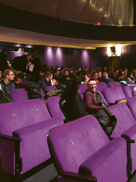 Ein Theater mit violetten Sitzen ist mit einem bunt gemischten Publikum gefüllt. Die Leute sitzen und unterhalten sich, während einige weiter hinten stehen. Die Kulisse ist gedämpft beleuchtet, was eine gemütliche Atmosphäre schafft.