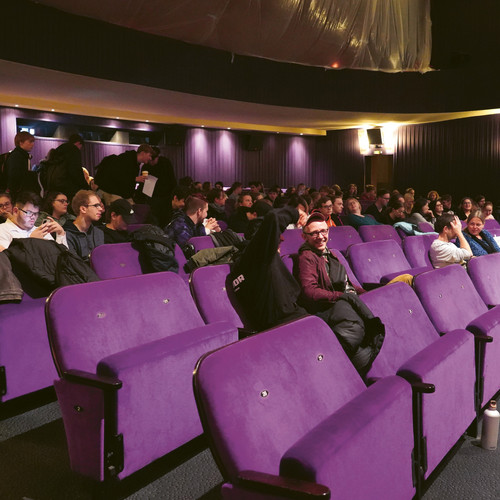 Ein Theater mit violetten Sitzen ist mit einem bunt gemischten Publikum gefüllt. Die Leute sitzen und unterhalten sich, während einige weiter hinten stehen. Die Kulisse ist gedämpft beleuchtet, was eine gemütliche Atmosphäre schafft.