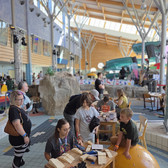 People of different ages gather in a bright, modern interior with large windows. Children and adults play at a table with wooden toys and building blocks. Colourful slides and rocks can be seen in the background.