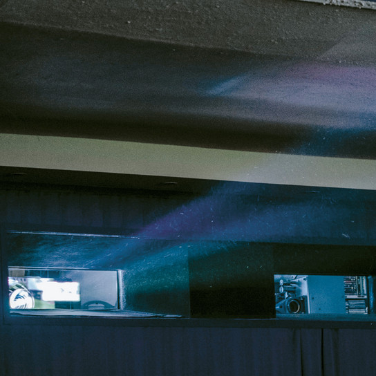 Ein Projektorstrahl fällt durch ein kleines Fenster in einen schwach beleuchteten Raum und verbreitet blaues und violettes Licht. Der Innenraum zeichnet sich durch niedrige Decken und dunkle Wände aus, was eine stimmungsvolle Atmosphäre schafft.