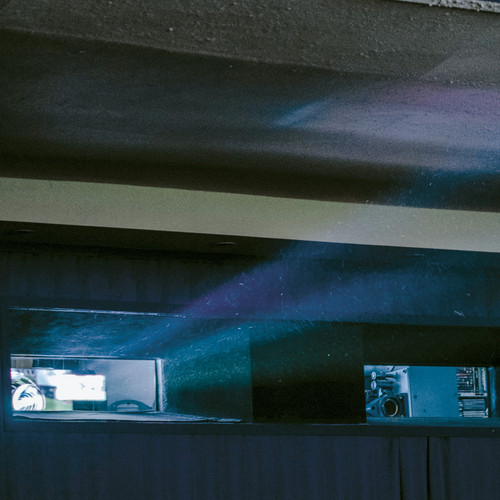 Ein Projektorstrahl fällt durch ein kleines Fenster in einen schwach beleuchteten Raum und verbreitet blaues und violettes Licht. Der Innenraum zeichnet sich durch niedrige Decken und dunkle Wände aus, was eine stimmungsvolle Atmosphäre schafft.
