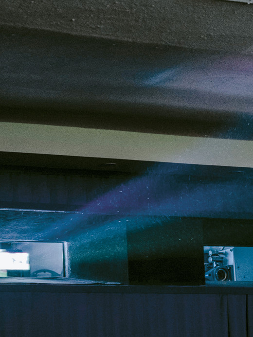 Ein Projektorstrahl fällt durch ein kleines Fenster in einen schwach beleuchteten Raum und verbreitet blaues und violettes Licht. Der Innenraum zeichnet sich durch niedrige Decken und dunkle Wände aus, was eine stimmungsvolle Atmosphäre schafft.