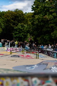 A skateboarder performs a trick on a rail in a graffiti-covered skate park while a crowd of spectators watches in the background, with trees and a partly cloudy sky in the background.