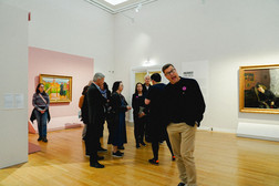 A group of people are standing in an art gallery, some of them looking at the paintings on the walls. A man in the foreground smiles and leans towards the camera. The room has a wooden floor and white and pink walls.