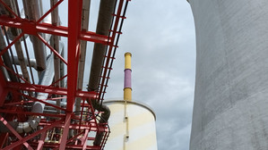 Ein gelb-violetter Schornstein steht zwischen zwei großen, grauen Industriekühltürmen. Rote Metallrohre und -träger kreuzen die linke Seite unter einem bewölkten Himmel.