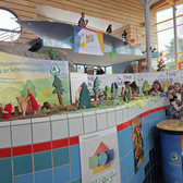 A colourful display of wooden animal and tree figures is set up on a tiled counter at an indoor event. People can be seen on an upper level, and banners and signs decorate the background.
