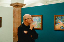 A bald man with glasses and a black shirt stands in an art gallery and contemplates the colourful paintings on a turquoise wall.