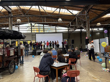 People sit at tables in a large industrial hall and watch a group of presenters standing on stage in front of a screen. Colourful balloons and decorations are scattered around the room.