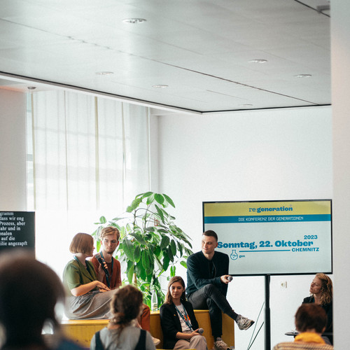 A small group of people are participating in a discussion in a modern conference room. Four speakers are seated at the front, facing an audience. A presentation slide on a screen shows the date, "Sonntag, 22. Oktober," and other German text. Colorful chairs are in the audience.