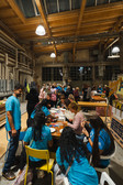 A large group of people, many in blue shirts, are gathered around tables inside, working on craft and art projects under a high wooden ceiling in a well-lit, industrial-looking room.