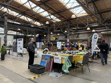 A creative fair is taking place in a large industrial-style hall, with colourful works of art and handicrafts on display on the tables. People browse the stalls and decorations hang from the ceiling. Bright light shines through the skylights in the wooden roof.