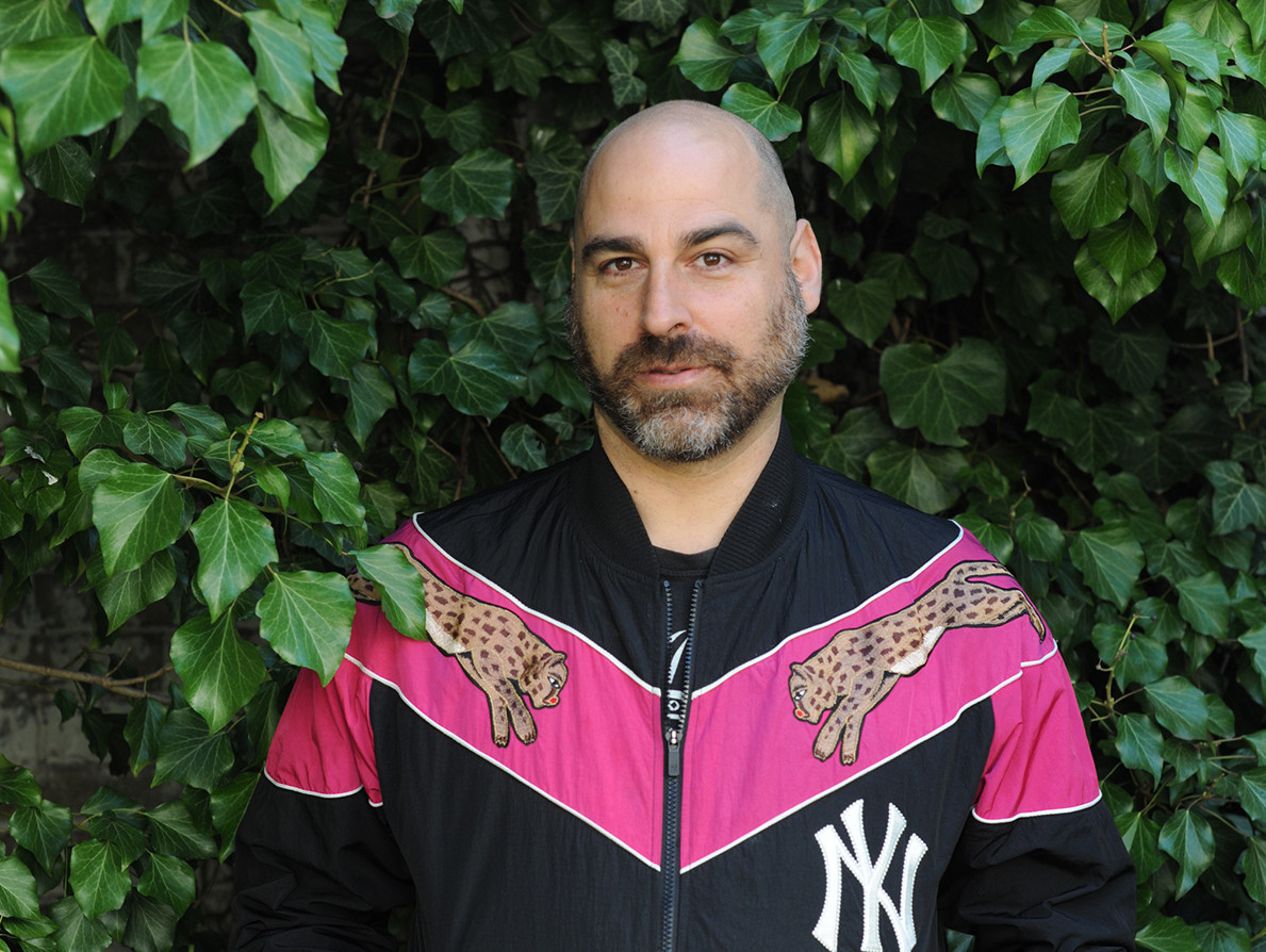 A person with a bald head and beard stands in front of a green background. She is wearing a black jacket with pink and magenta-coloured accents, cheetah motifs and a New York Yankees logo. Her hands are in the pockets of her jacket.