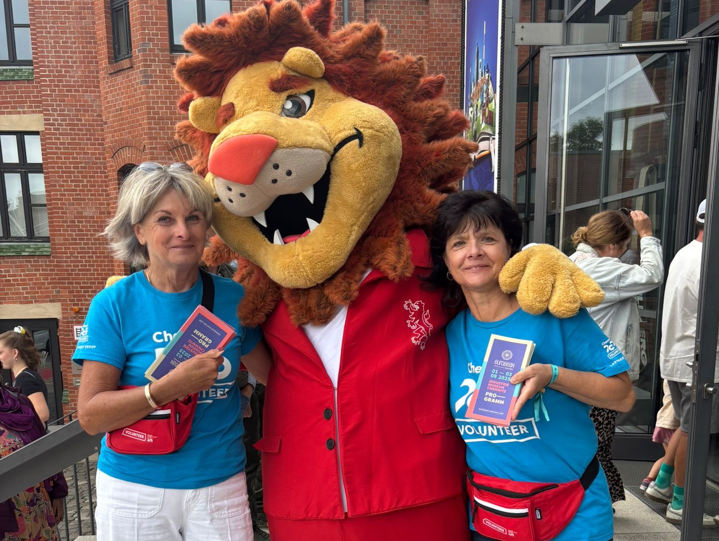 In front of a brick building with a glass entrance, two women in blue volunteer shirts stand next to a person in a lion mascot costume, all smiling and holding event brochures.