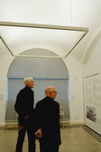 Two men with short hair and dark clothes stand in a minimalist gallery space and look at an exhibition text on a white wall in soft daylight.