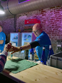 A man in a blue shirt stands behind a bar and hands a bottled drink to a customer. The bar has glass bottles, a green cloth and fridges with drinks in the background in front of an exposed brick wall.