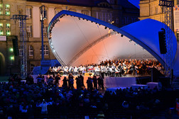 Ein großes Orchester spielt auf einer hell erleuchteten Freilichtbühne mit einem modernen, geschwungenen Baldachin bei Nacht. Das Publikum sitzt in Stuhlreihen, und Fotografen stehen in der Nähe der Bühne und halten das Geschehen fest.