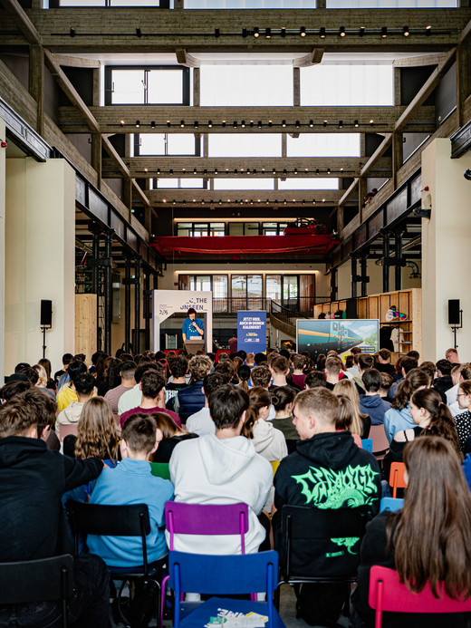 A large group of people is seated and facing a stage in an industrial-style hall with high ceilings and exposed beams. The audience is diverse with many young individuals. On stage, a person is speaking in front of a screen and banners.