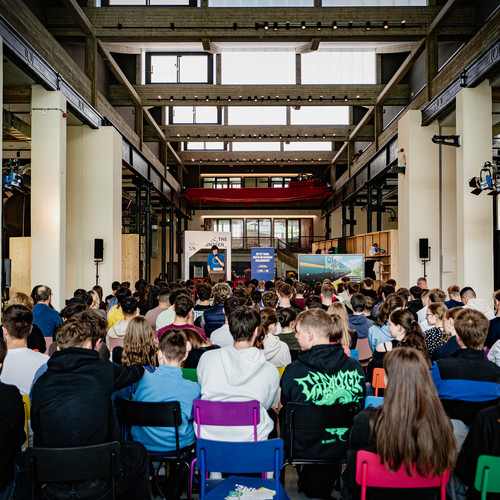 A large group of people is seated and facing a stage in an industrial-style hall with high ceilings and exposed beams. The audience is diverse with many young individuals. On stage, a person is speaking in front of a screen and banners.