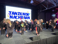 A group of older women in black clothing perform a synchronised dance on stage, bending forwards with their arms outstretched. Behind them, the words "DANCING NEIGHBOURS" appear on a screen on a colourful background.