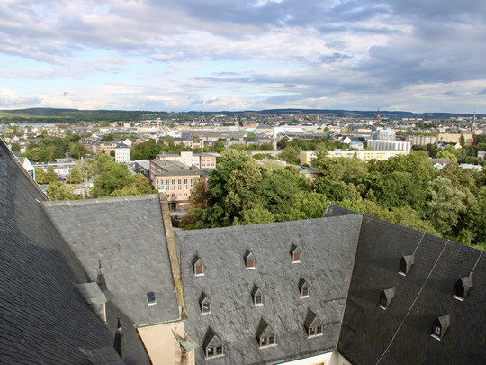 A view of the city from above shows slate-covered roofs with triangular windows, green trees and a sprawling city stretching into the distance under a partly cloudy sky.