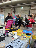 Three people stand at a craft table with paint and stencils and print carrier bags in an industrial indoor environment. Rolls of adhesive tape, paint and art materials are spread out on the table.