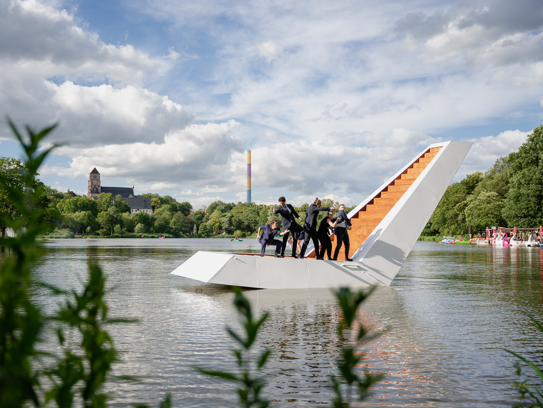 Eine Gruppe von Menschen in schwarzen Anzügen steht auf einer ungewöhnlichen schwimmenden Struktur mit einer steilen, orangefarbenen Treppe inmitten eines ruhigen Sees, umgeben von Grün. Im Hintergrund sind unter einem teilweise bewölkten Himmel eine Kirche und ein hoher, gestreifter Schornstein zu sehen.